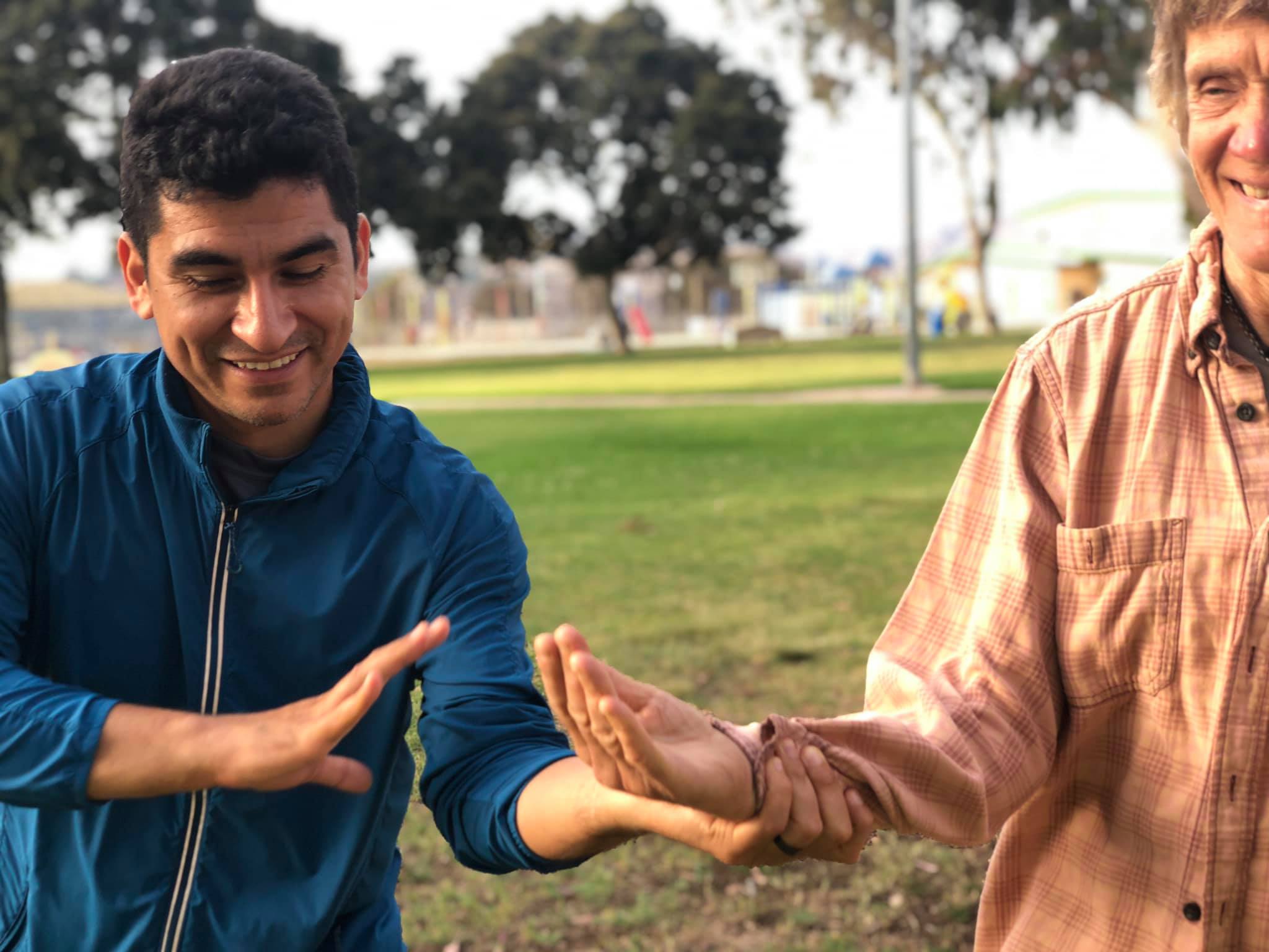 Dojomates Marco and Rosalind training together in the park on a sunny morning. Marco has grabbed Rosalind's wrist and is moving without resistance as she turns in tai no henko. Both are smiling.