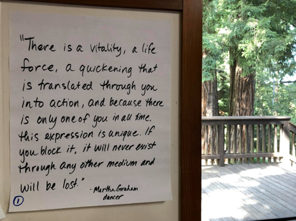 A photo of a large, handwritten quote, in black marker, posted on the wall at an Aikido workshop in the redwoods. Trees are visible through the window next to the paper. The quote reads: "There is a vitality, a life force, a quickening 
that is translated through you into action, 
and there is only one of you in all time, 
this expression is unique, 
and if you block it, 
it will never exist through any other medium; and be lost. 
Martha Graham, dancer"