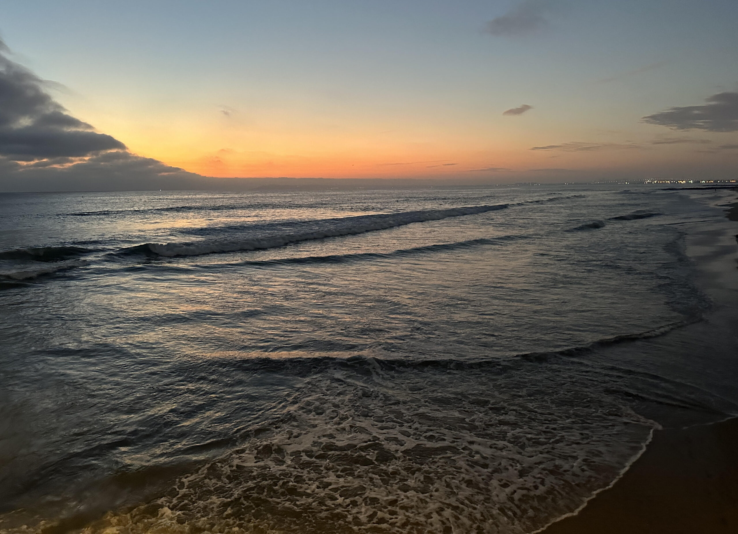 A photo of ocean waves breaking and rolling in at Imperial Beach just after sunset, with city lights and a few clouds in the distance.